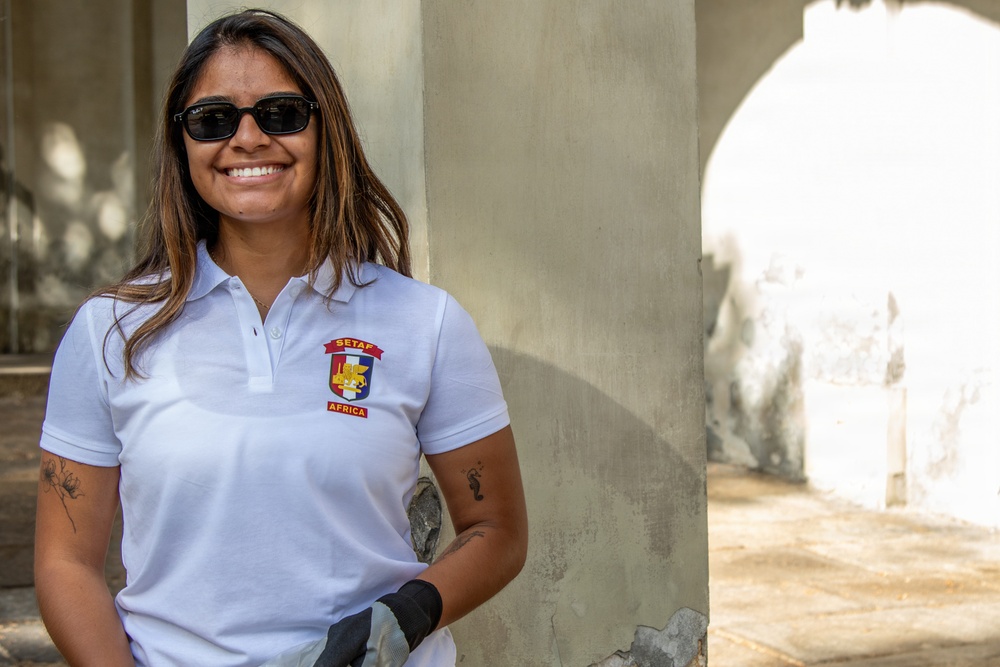 A smiling woman wearing a white polo shirt with a 'SETAF Africa' emblem stands leaning against a concrete pillar. She is wearing dark sunglasses and black work gloves, suggesting she is engaged in some form of volunteer or community service activity. The setting appears to be an outdoor or semi-covered structure with weathered concrete walls.