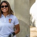 A smiling woman wearing a white polo shirt with a 'SETAF Africa' emblem stands leaning against a concrete pillar. She is wearing dark sunglasses and black work gloves, suggesting she is engaged in some form of volunteer or community service activity. The setting appears to be an outdoor or semi-covered structure with weathered concrete walls.