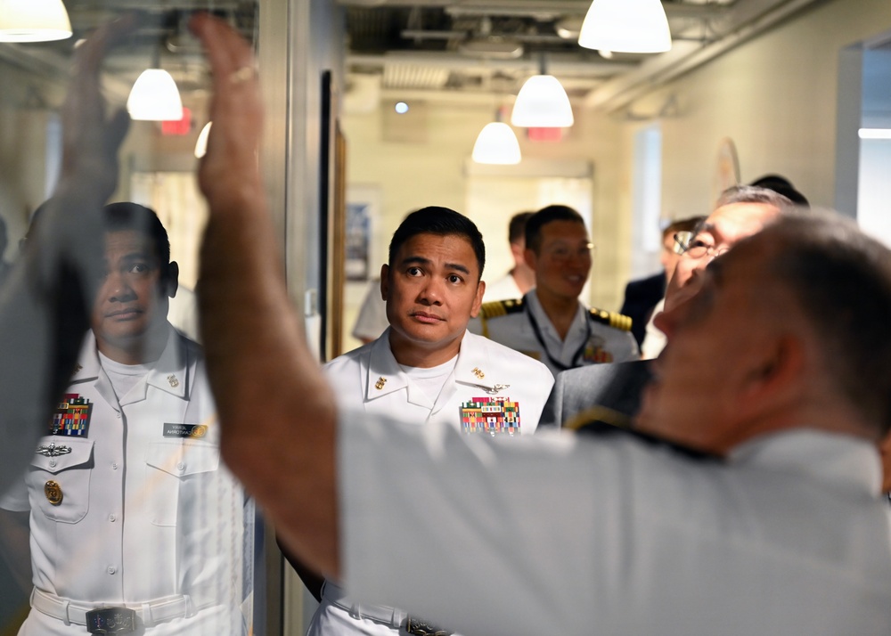 A group of naval officers in white dress uniforms are gathered in what appears to be an indoor facility, likely during an official tour or briefing. A person in the foreground has their arm raised, possibly pointing at or gesturing toward a glass partition or display. The officers are attentively observing something being presented or explained to them.