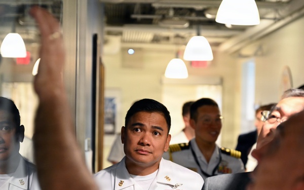 A group of naval officers in white dress uniforms are gathered in what appears to be an indoor facility, likely during an official tour or briefing. A person in the foreground has their arm raised, possibly pointing at or gesturing toward a glass partition or display. The officers are attentively observing something being presented or explained to them.
