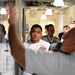 A group of naval officers in white dress uniforms are gathered in what appears to be an indoor facility, likely during an official tour or briefing. A person in the foreground has their arm raised, possibly pointing at or gesturing toward a glass partition or display. The officers are attentively observing something being presented or explained to them.