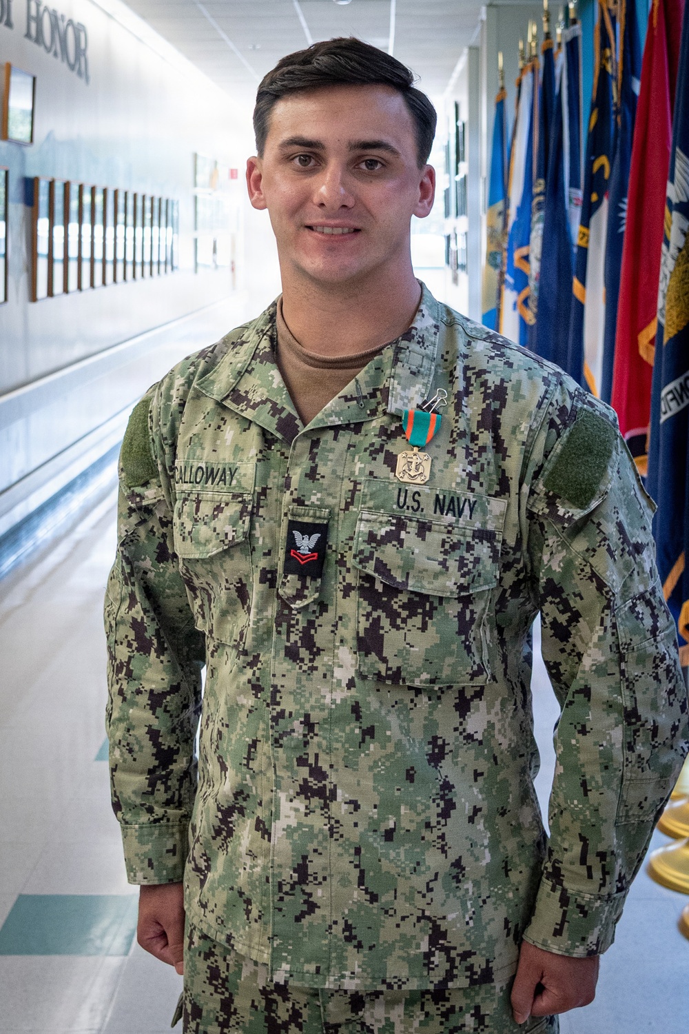 A U.S. Navy sailor in NWU Type I (digital camouflage) uniform is posing in a military hallway flanked by service flags. The sailor, identified by name tape as 'CALLOWAY,' is wearing a Petty Officer Third Class rank insignia and a service medal with a teal and orange ribbon. The setting appears to be an official military building, likely during or after an awards ceremony.