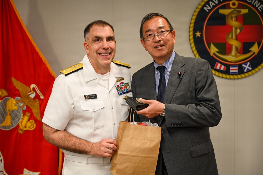 A U.S. Navy officer in white dress uniform, identified by name tag as 'Matthew Case', poses with a civilian man in a gray suit who is wearing what appears to be a Japanese Diet member's pin. The two individuals appear to be exchanging or presenting gifts, including a brown paper bag and a medallion or coin. The setting appears to be an official military ceremony or meeting, with a Navy Medicine emblem and U.S. Marine Corps flag visible in the background.