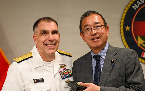 A U.S. Navy officer in white dress uniform, identified by name tag as 'Matthew Case', poses with a civilian man in a gray suit who is wearing what appears to be a Japanese Diet member's pin. The two individuals appear to be exchanging or presenting gifts, including a brown paper bag and a medallion or coin. The setting appears to be an official military ceremony or meeting, with a Navy Medicine emblem and U.S. Marine Corps flag visible in the background.