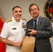 A U.S. Navy officer in white dress uniform, identified by name tag as 'Matthew Case', poses with a civilian man in a gray suit who is wearing what appears to be a Japanese Diet member's pin. The two individuals appear to be exchanging or presenting gifts, including a brown paper bag and a medallion or coin. The setting appears to be an official military ceremony or meeting, with a Navy Medicine emblem and U.S. Marine Corps flag visible in the background.