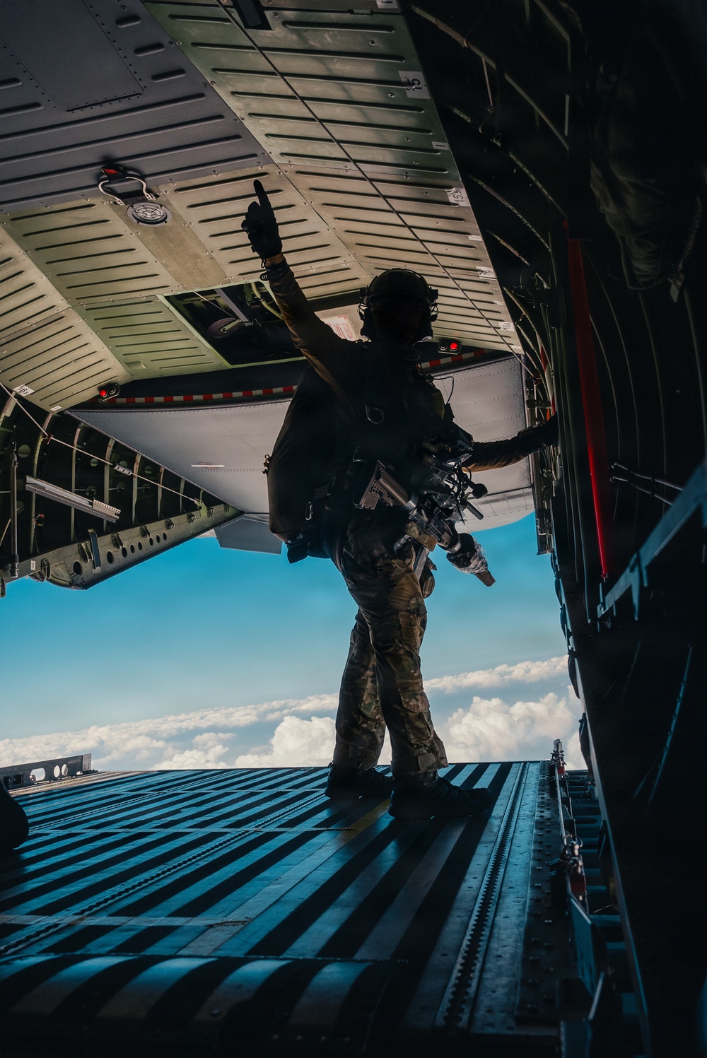 A fully equipped military personnel stands at the open rear ramp of a large military transport aircraft in flight, raising one hand in a signaling gesture. The individual is silhouetted against a bright blue sky with clouds visible below, indicating significant altitude. The scene appears to depict a pre-jump or cargo-drop preparation sequence.