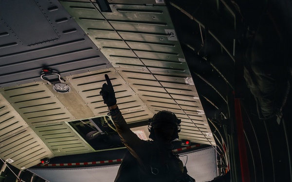 A fully equipped military personnel stands at the open rear ramp of a large military transport aircraft in flight, raising one hand in a signaling gesture. The individual is silhouetted against a bright blue sky with clouds visible below, indicating significant altitude. The scene appears to depict a pre-jump or cargo-drop preparation sequence.