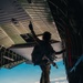 A fully equipped military personnel stands at the open rear ramp of a large military transport aircraft in flight, raising one hand in a signaling gesture. The individual is silhouetted against a bright blue sky with clouds visible below, indicating significant altitude. The scene appears to depict a pre-jump or cargo-drop preparation sequence.