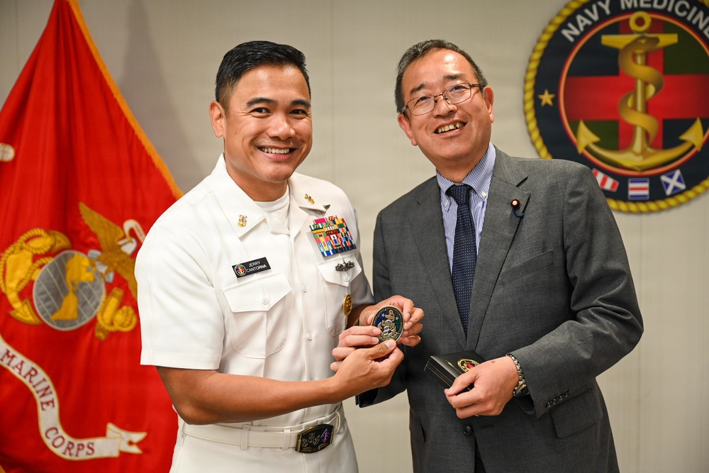 A U.S. Navy officer in dress white uniform, identified by nameplate as 'Jerry Canturna,' is presenting a challenge coin to a civilian man in a gray suit. The ceremony takes place in front of a U.S. Marine Corps flag and a Navy Medicine organizational seal. Both individuals are smiling, suggesting a formal but positive diplomatic or ceremonial exchange.