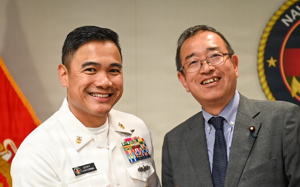 A U.S. Navy officer in dress white uniform, identified by nameplate as 'Jerry Canturna,' is presenting a challenge coin to a civilian man in a gray suit. The ceremony takes place in front of a U.S. Marine Corps flag and a Navy Medicine organizational seal. Both individuals are smiling, suggesting a formal but positive diplomatic or ceremonial exchange.
