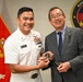 A U.S. Navy officer in dress white uniform, identified by nameplate as 'Jerry Canturna,' is presenting a challenge coin to a civilian man in a gray suit. The ceremony takes place in front of a U.S. Marine Corps flag and a Navy Medicine organizational seal. Both individuals are smiling, suggesting a formal but positive diplomatic or ceremonial exchange.