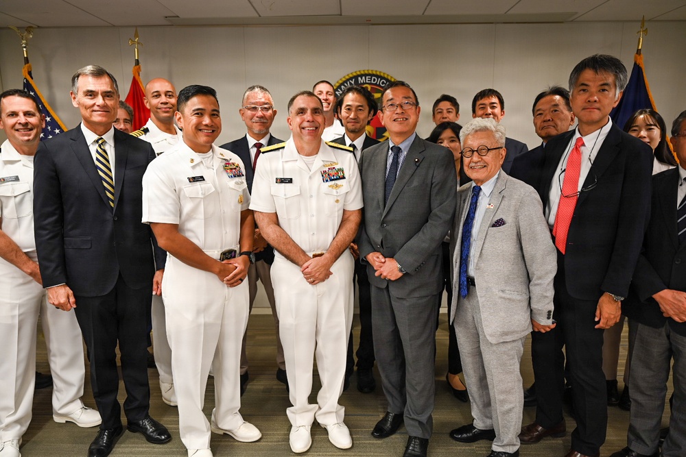 A formal group photo taken at a Navy Medicine facility, showing U.S. Navy personnel in white dress uniforms alongside civilians in business attire, including what appears to be a Japanese delegation. The setting features American and other flags in the background along with a 'Navy Medicine' seal/logo on the wall. The gathering suggests an official diplomatic or military-civilian exchange event.