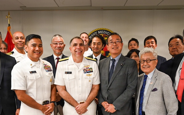 A formal group photo taken at a Navy Medicine facility, showing U.S. Navy personnel in white dress uniforms alongside civilians in business attire, including what appears to be a Japanese delegation. The setting features American and other flags in the background along with a 'Navy Medicine' seal/logo on the wall. The gathering suggests an official diplomatic or military-civilian exchange event.