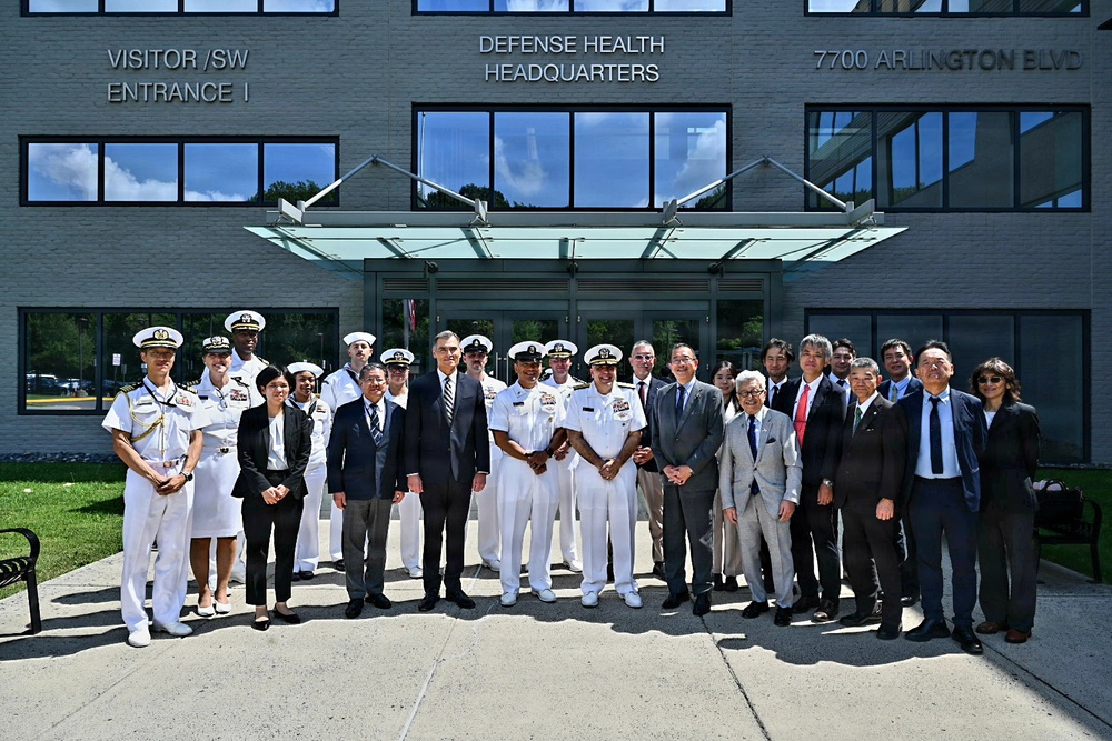 A group of approximately 20 individuals poses for a formal group photograph in front of the Defense Health Headquarters building located at 7700 Arlington Blvd. The group includes U.S. Navy personnel in white dress uniforms and civilians in business attire, suggesting an official diplomatic or professional meeting. The mix of individuals appears to include both American military/civilian officials and what appear to be Japanese or Asian civilian delegates.