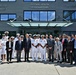 A group of approximately 20 individuals poses for a formal group photograph in front of the Defense Health Headquarters building located at 7700 Arlington Blvd. The group includes U.S. Navy personnel in white dress uniforms and civilians in business attire, suggesting an official diplomatic or professional meeting. The mix of individuals appears to include both American military/civilian officials and what appear to be Japanese or Asian civilian delegates.