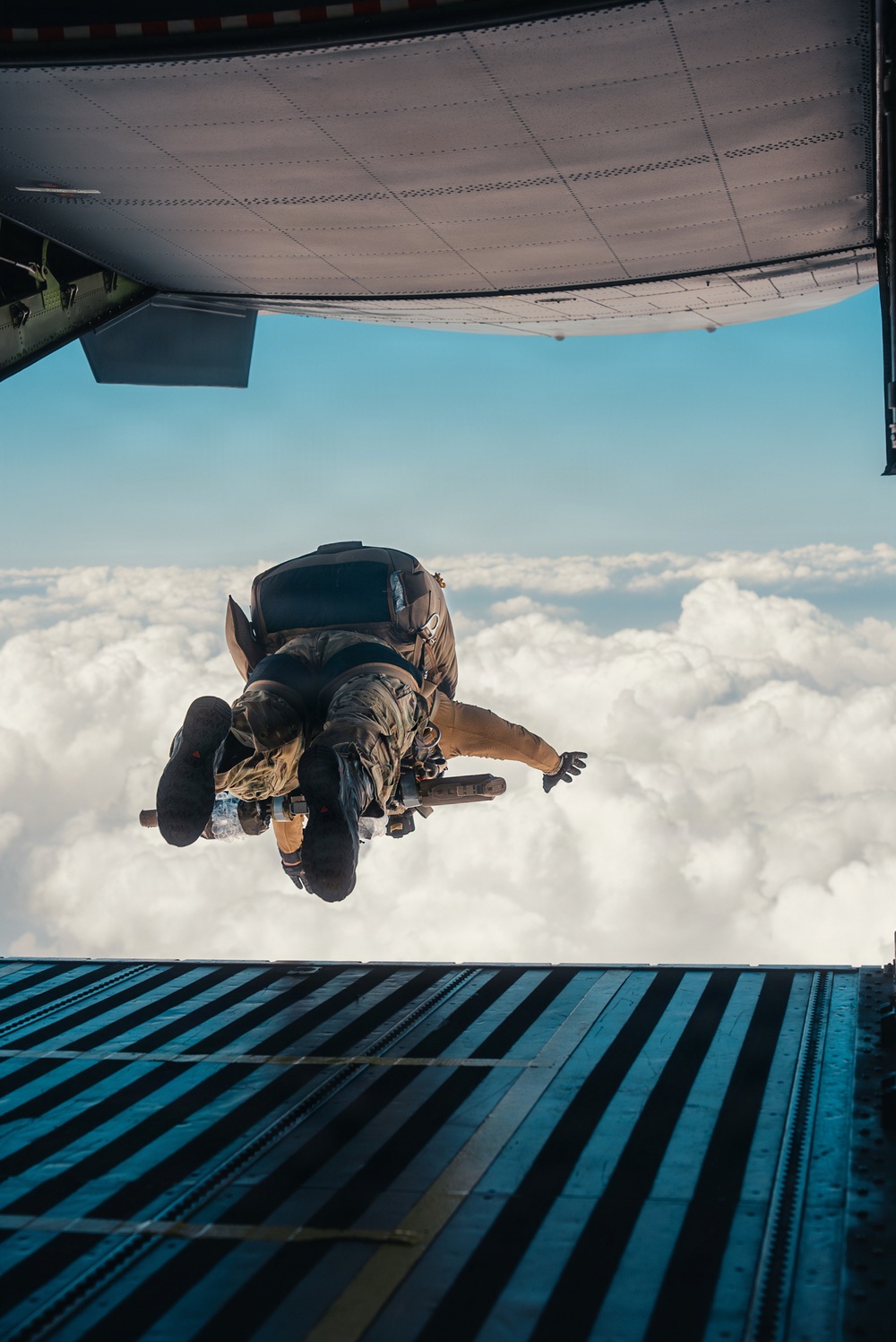 Two individuals are captured mid-exit from the open rear ramp of a military cargo aircraft, appearing to be in a tandem skydive configuration above a cloud layer. The photo is taken from inside the aircraft looking outward, showing the jumpers just moments after leaving the ramp. The scene suggests a military or tactical parachute operation given the equipment and aircraft type.