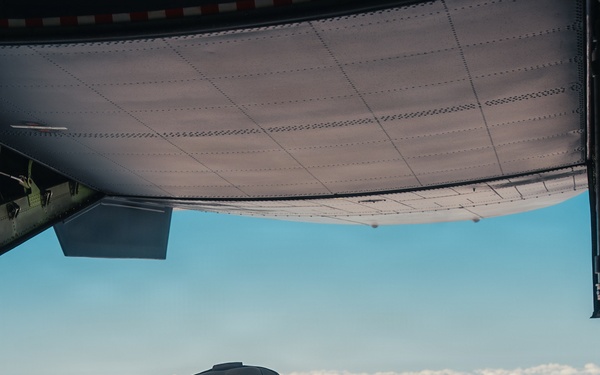 Two individuals are captured mid-exit from the open rear ramp of a military cargo aircraft, appearing to be in a tandem skydive configuration above a cloud layer. The photo is taken from inside the aircraft looking outward, showing the jumpers just moments after leaving the ramp. The scene suggests a military or tactical parachute operation given the equipment and aircraft type.