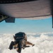 Two individuals are captured mid-exit from the open rear ramp of a military cargo aircraft, appearing to be in a tandem skydive configuration above a cloud layer. The photo is taken from inside the aircraft looking outward, showing the jumpers just moments after leaving the ramp. The scene suggests a military or tactical parachute operation given the equipment and aircraft type.