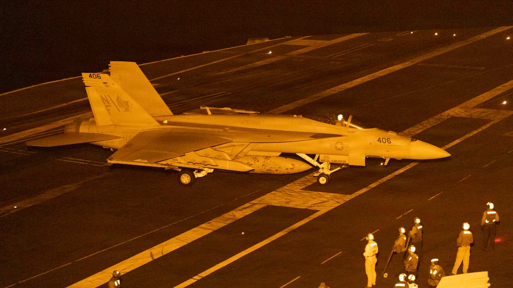 A U.S. Navy F/A-18 Super Hornet (Bureau Number 406) is positioned on an aircraft carrier flight deck at night, illuminated by orange/amber artificial lighting. The aircraft displays a squadron insignia on its tail and is surrounded by flight deck personnel in various colored jerseys. The scene depicts a nighttime carrier flight deck operation, likely pre-launch or post-recovery.