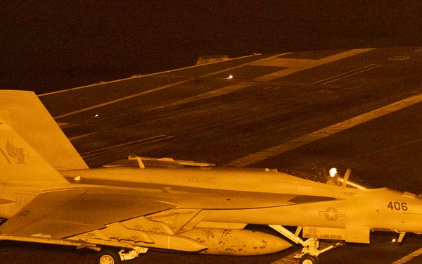 A U.S. Navy F/A-18 Super Hornet (Bureau Number 406) is positioned on an aircraft carrier flight deck at night, illuminated by orange/amber artificial lighting. The aircraft displays a squadron insignia on its tail and is surrounded by flight deck crew members in various colored jerseys. The scene depicts a nighttime carrier flight deck operation, likely pre-launch or post-recovery.