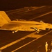 A U.S. Navy F/A-18 Super Hornet (Bureau Number 406) is positioned on an aircraft carrier flight deck at night, illuminated by orange/amber artificial lighting. The aircraft displays a squadron insignia on its tail and is surrounded by flight deck personnel in various colored jerseys. The scene depicts a nighttime carrier flight deck operation, likely pre-launch or post-recovery.