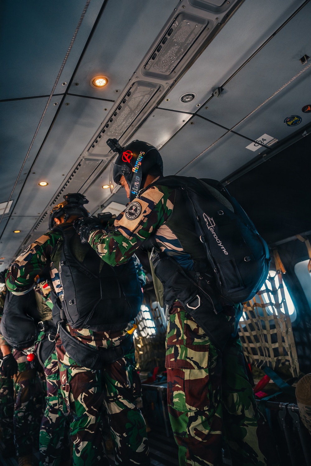 Military paratroopers are inside a transport aircraft preparing for a jump operation. Personnel are wearing camouflage uniforms with parachute rigs, helmets, and tactical gear. The scene depicts a pre-jump equipment check or buddy check routine aboard the aircraft.
