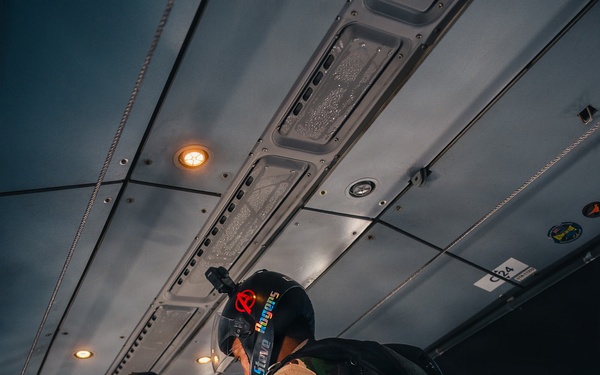 Military paratroopers are inside a transport aircraft preparing for a jump operation. Personnel are wearing camouflage uniforms with parachute rigs, helmets, and tactical gear. The scene depicts a pre-jump equipment check or buddy check routine aboard the aircraft.