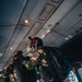 Military paratroopers are inside a transport aircraft preparing for a jump operation. Personnel are wearing camouflage uniforms with parachute rigs, helmets, and tactical gear. The scene depicts a pre-jump equipment check or buddy check routine aboard the aircraft.