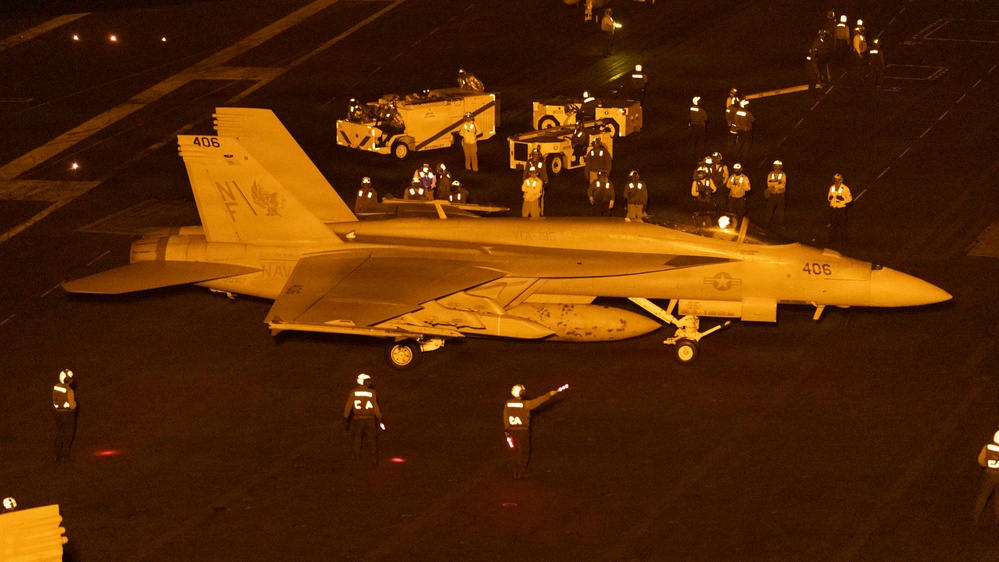 A U.S. Navy F/A-18 Hornet or Super Hornet (modex 406) is positioned on an aircraft carrier flight deck at night, illuminated by orange/amber lighting. Numerous flight deck crew members in color-coded jerseys and helmets surround the aircraft, along with ground support vehicles. The scene depicts a typical night flight deck operation aboard a U.S. Navy carrier.