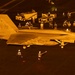 A U.S. Navy F/A-18 Hornet or Super Hornet (modex 406) is positioned on an aircraft carrier flight deck at night, illuminated by orange/amber lighting. Numerous flight deck crew members in color-coded jerseys and helmets surround the aircraft, along with ground support vehicles. The scene depicts a typical night flight deck operation aboard a U.S. Navy carrier.