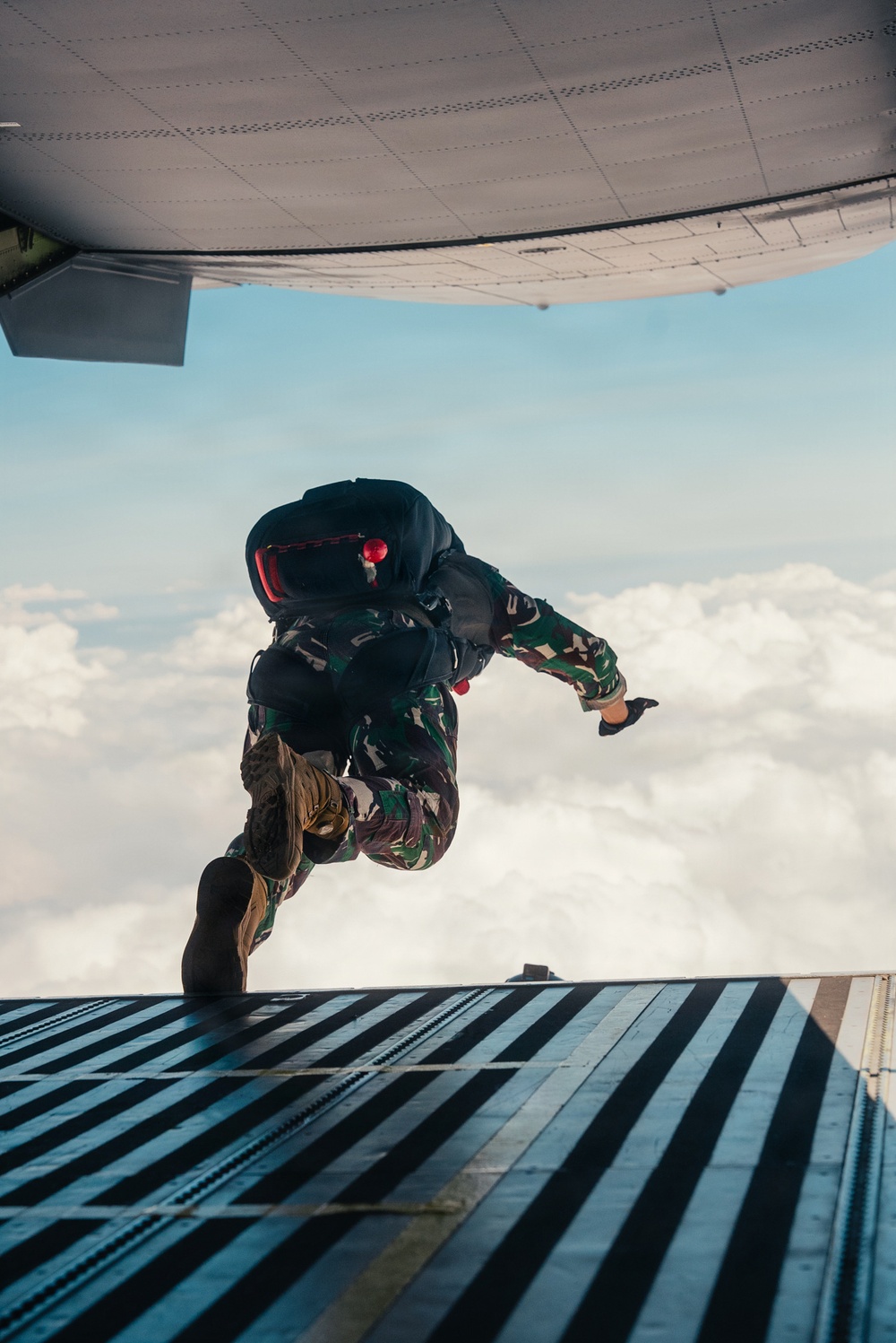 A military paratrooper in camouflage uniform is captured mid-jump exiting the rear ramp of a military transport aircraft at high altitude. The jumper is wearing a full parachute rig with a reserve chute and tactical gear. The image is taken from inside the aircraft looking outward, with clouds visible in the background below.
