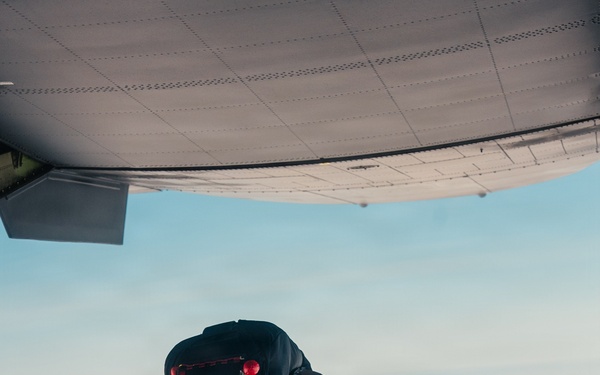 A military paratrooper in camouflage uniform is captured mid-jump exiting the rear ramp of a military transport aircraft at high altitude. The jumper is wearing a full parachute rig with a reserve chute and tactical gear. The image is taken from inside the aircraft looking outward, with clouds visible in the background below.