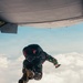 A military paratrooper in camouflage uniform is captured mid-jump exiting the rear ramp of a military transport aircraft at high altitude. The jumper is wearing a full parachute rig with a reserve chute and tactical gear. The image is taken from inside the aircraft looking outward, with clouds visible in the background below.