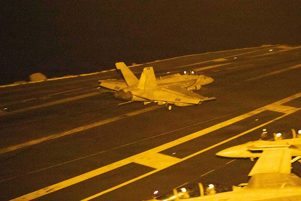 A nighttime carrier deck scene showing at least two military jet aircraft, with the foreground aircraft marked '106' appearing to be in motion on the flight deck. The image is bathed in amber/orange lighting typical of carrier deck night operations. A third aircraft is partially visible in the lower right corner of the frame.