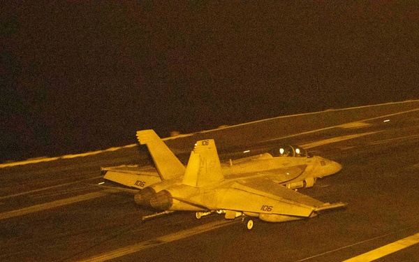 A nighttime carrier deck scene showing at least two military jet aircraft, with the foreground aircraft marked '106' appearing to be in motion on the flight deck. The image is bathed in amber/orange lighting typical of carrier deck night operations. A third aircraft is partially visible in the lower right corner of the frame.