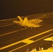 A nighttime carrier deck scene showing at least two military jet aircraft, with the foreground aircraft marked '106' appearing to be in motion on the flight deck. The image is bathed in amber/orange lighting typical of carrier deck night operations. A third aircraft is partially visible in the lower right corner of the frame.