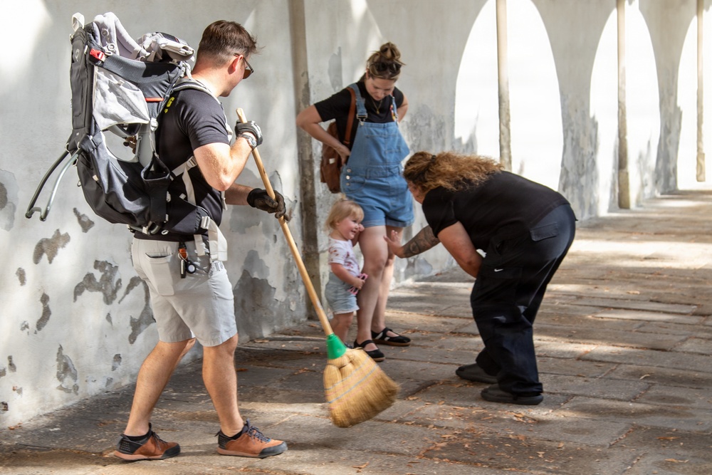 A group of people are gathered in an outdoor covered walkway or arcade with white arched walls. A man wearing a large backpack and gloves is sweeping the stone-paved floor with a broom, while a person in black bends down toward a small toddler. A woman in denim overalls stands nearby watching the child.