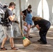 A group of people are gathered in an outdoor covered walkway or arcade with white arched walls. A man wearing a large backpack and gloves is sweeping the stone-paved floor with a broom, while a person in black bends down toward a small toddler. A woman in denim overalls stands nearby watching the child.