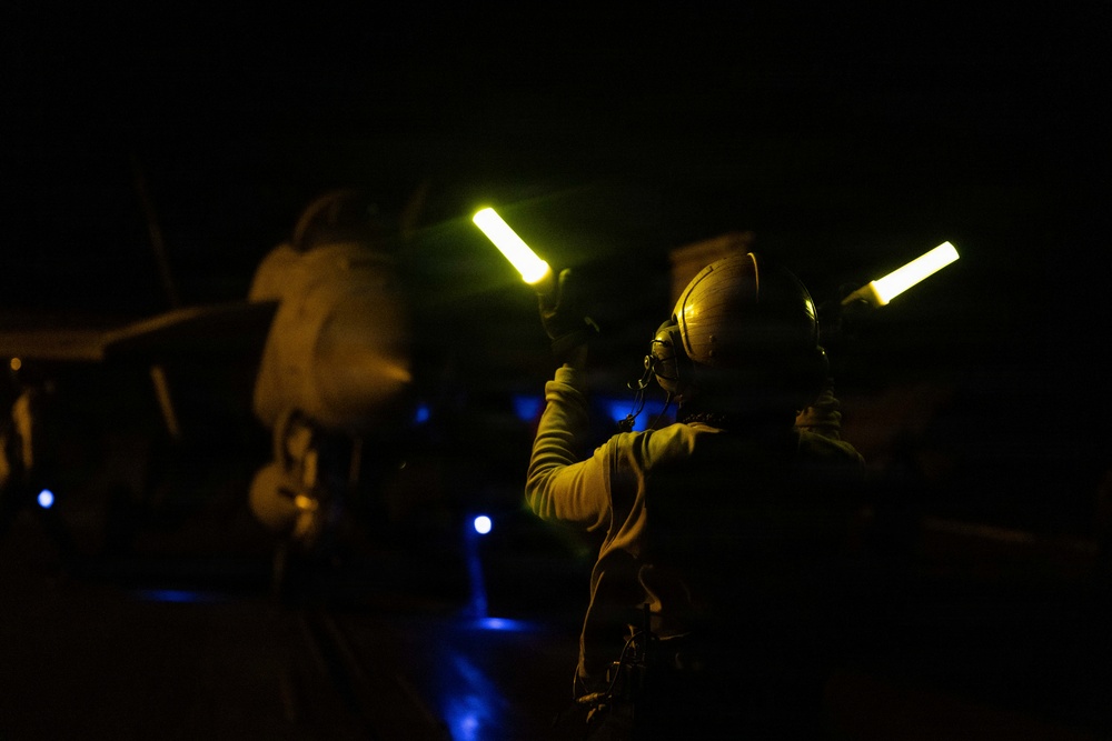 A night operations scene on what appears to be a military aircraft carrier flight deck. A deck crew member wearing a flight helmet and hearing protection is using illuminated wands (glow sticks or light wands) to signal or marshal an aircraft during nighttime operations. Blue deck lighting and the silhouette of a military aircraft are visible in the background.