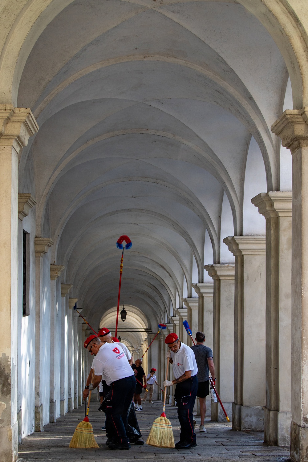 A group of workers in matching uniforms — white shirts with a red cross/maltese cross logo and red berets — are sweeping a grand colonnaded arcade with traditional broom and long-handled brushes. The architectural setting features a long barrel-vaulted corridor with repeating stone arches receding into the distance, suggesting a historic European building, likely in Italy. The scene captures an organized cleaning operation in a monumental public space.