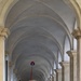 A group of workers in matching uniforms — white shirts with a red cross/maltese cross logo and red berets — are sweeping a grand colonnaded arcade with traditional broom and long-handled brushes. The architectural setting features a long barrel-vaulted corridor with repeating stone arches receding into the distance, suggesting a historic European building, likely in Italy. The scene captures an organized cleaning operation in a monumental public space.
