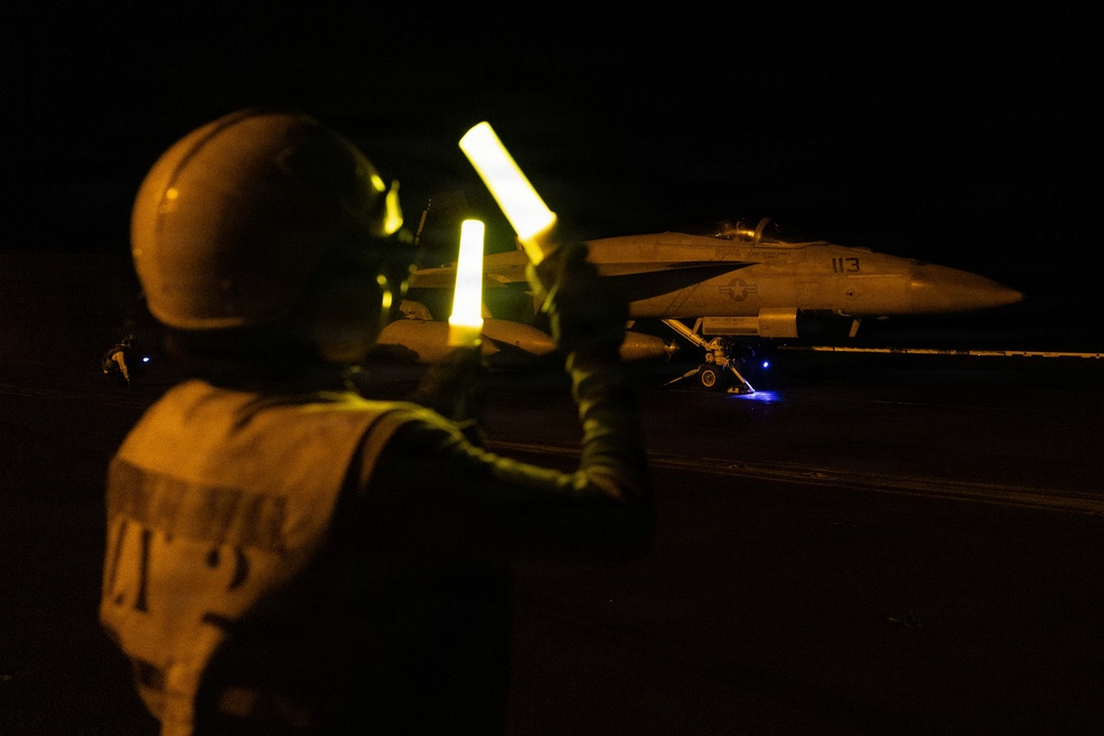 A night flight deck operation aboard what appears to be a U.S. Navy aircraft carrier. A plane director (yellowshirt) is using illuminated wands to marshal a military jet aircraft, numbered '113', on the darkened flight deck. The scene is lit primarily by the glow of the wands and ambient deck lighting.