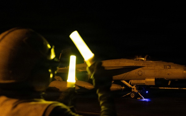 A night flight deck operation aboard what appears to be a U.S. Navy aircraft carrier. A plane director (yellowshirt) is using illuminated wands to marshal a military jet aircraft, numbered '113', on the darkened flight deck. The scene is lit primarily by the glow of the wands and ambient deck lighting.