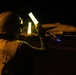 A night flight deck operation aboard what appears to be a U.S. Navy aircraft carrier. A plane director (yellowshirt) is using illuminated wands to marshal a military jet aircraft, numbered '113', on the darkened flight deck. The scene is lit primarily by the glow of the wands and ambient deck lighting.