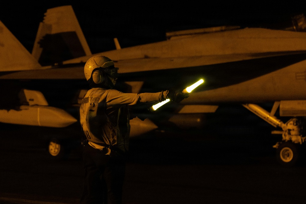 A naval flight deck crew member is directing aircraft during nighttime flight operations, holding an illuminated wand (light baton) used for signaling. The scene takes place on what appears to be an aircraft carrier flight deck, with a military jet aircraft — likely an F/A-18 Hornet or Super Hornet — visible in the background. The low-light conditions are characteristic of night carrier operations.