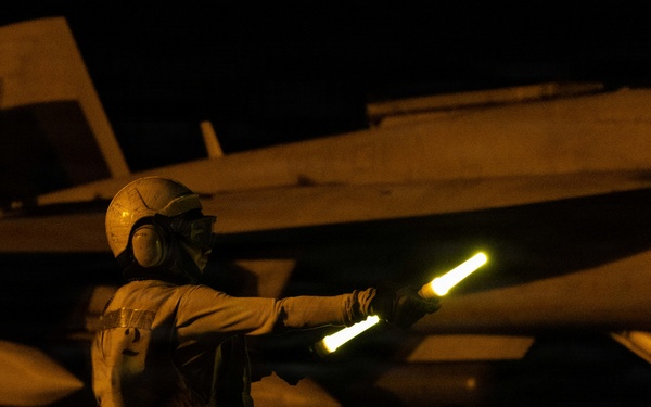 A naval flight deck crew member is directing aircraft during nighttime flight operations, holding an illuminated wand (light baton) used for signaling. The scene takes place on what appears to be an aircraft carrier flight deck, with a military jet aircraft — likely an F/A-18 Hornet or Super Hornet — visible in the background. The low-light conditions are characteristic of night carrier operations.