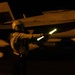 A naval flight deck crew member is directing aircraft during nighttime flight operations, holding an illuminated wand (light baton) used for signaling. The scene takes place on what appears to be an aircraft carrier flight deck, with a military jet aircraft — likely an F/A-18 Hornet or Super Hornet — visible in the background. The low-light conditions are characteristic of night carrier operations.