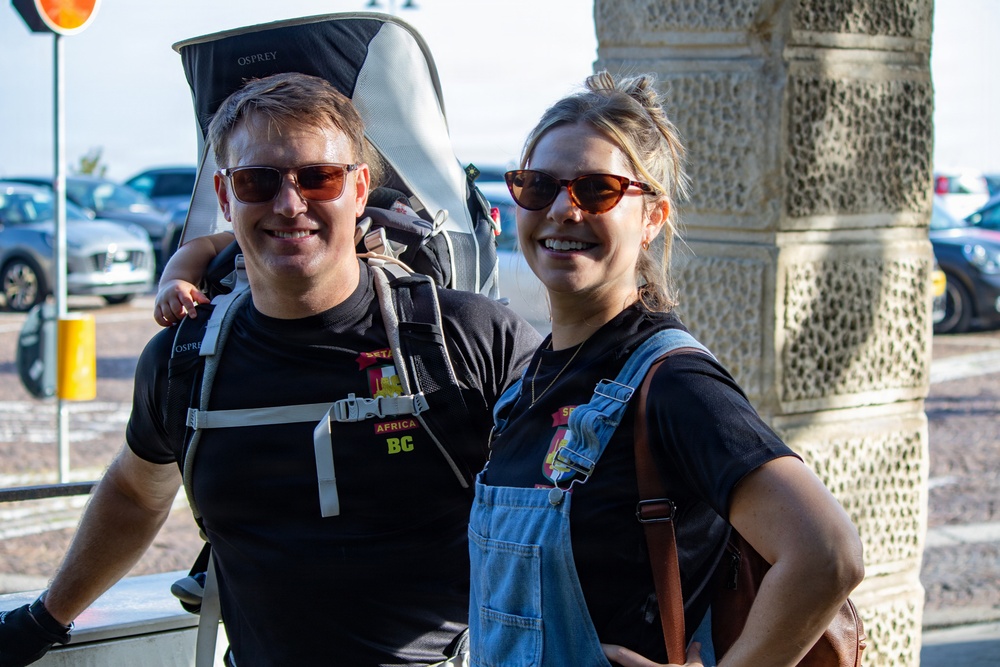 Two smiling adults pose together outdoors, both wearing sunglasses and matching black t-shirts with colorful badges/patches. The man on the left is carrying a large Osprey brand child carrier backpack. They appear to be travelers or participants in some kind of organized event, standing near a decorative stone pillar with a parking lot visible in the background.