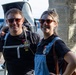 Two smiling adults pose together outdoors, both wearing sunglasses and matching black t-shirts with colorful badges/patches. The man on the left is carrying a large Osprey brand child carrier backpack. They appear to be travelers or participants in some kind of organized event, standing near a decorative stone pillar with a parking lot visible in the background.