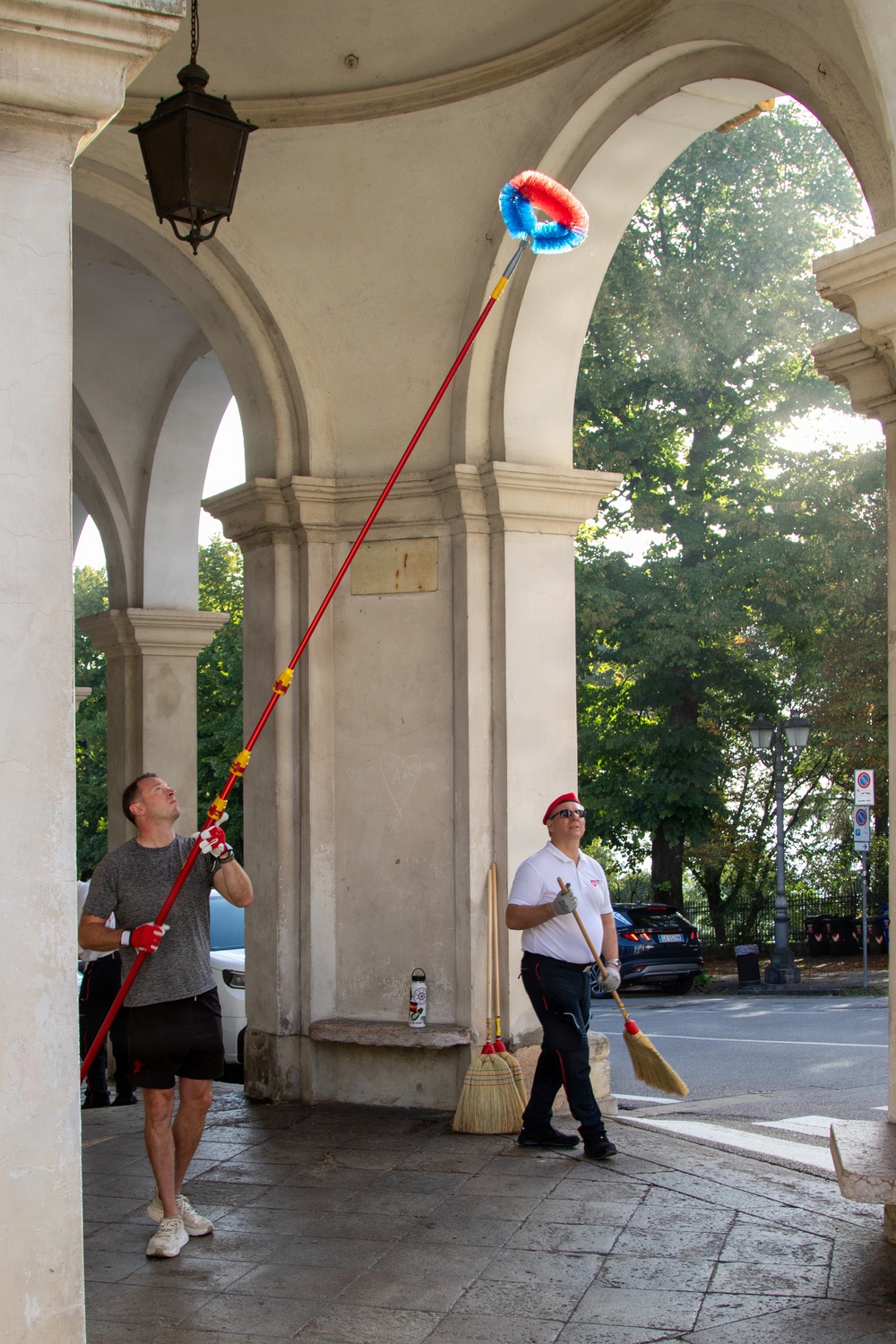 Two workers are cleaning the vaulted ceiling and floor of a classical arcade or portico with white arched columns. One worker uses a long red telescoping pole with a colorful blue-and-red duster head to reach the upper ceiling, while the other holds a traditional broom. The setting appears to be an Italian public building or monument with elegant architectural arches.